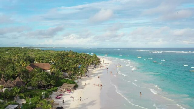 Vista a&eacute;rea panor&aacute;mica de Playa Para&iacute;so en Tulum, el drone descendiendo lentamente con muchos turistas caminando sobre la playa y nadando en el mar. Un cielo azul con nubes como fondo.