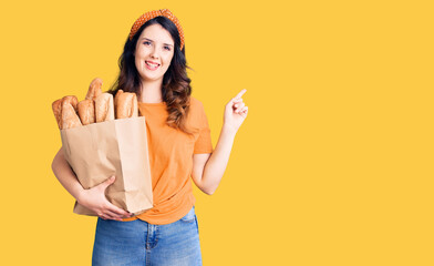 Beautiful young brunette woman holding paper bag with bread smiling happy pointing with hand and finger to the side