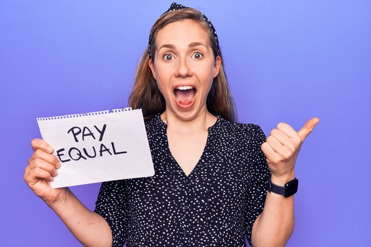 Young beautiful blonde woman holding paper with pay equal protest text pointing thumb up to the side smiling happy with open mouth