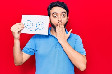 Young hispanic man holding sad to happy emotion paper covering mouth with hand, shocked and afraid...