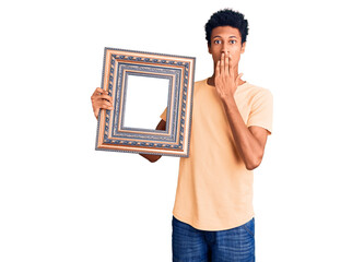 Young african american man holding empty frame covering mouth with hand, shocked and afraid for...