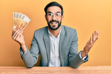 Young hispanic man holding norwegian krone banknotes sitting on the table celebrating achievement...