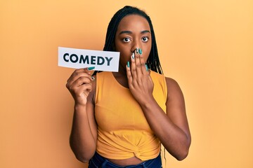 Young african american woman holding comedy word paper covering mouth with hand, shocked and afraid...