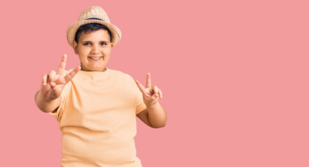 Little boy kid wearing summer hat and hawaiian swimsuit smiling looking to the camera showing fingers doing victory sign. number two.