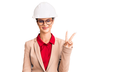 Young beautiful woman wearing architect hardhat smiling with happy face winking at the camera doing victory sign. number two.