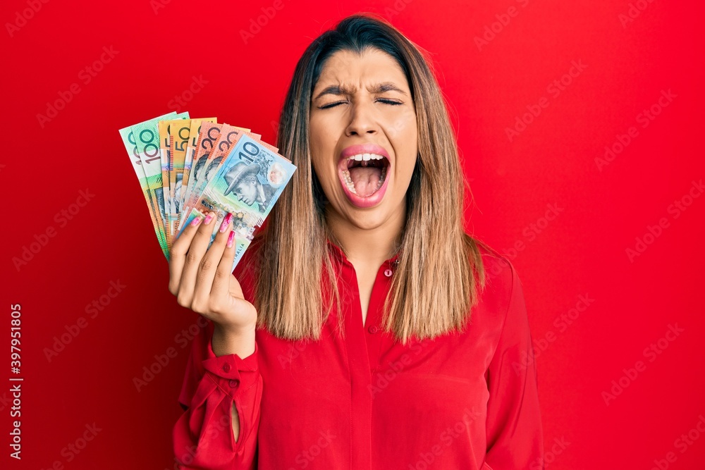 Poster beautiful brunette woman holding australian dollars angry and mad screaming frustrated and furious, 