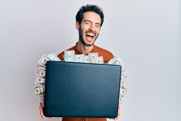 Young hispanic man holding briefcase with dollars smiling and laughing hard out loud because funny...