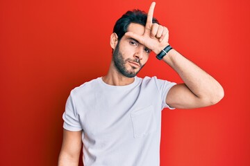 Young hispanic man wearing casual white tshirt making fun of people with fingers on forehead doing...
