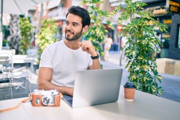 Young hispanic man smiling happy working using laptop at coffee shop terrace.