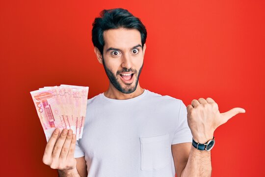Young hispanic man holding yuan chinese banknotes pointing thumb up to the side smiling happy with open mouth