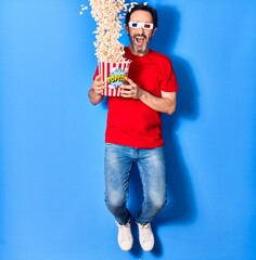 Middle age handsome hispanic man watching movie using 3d glasses similing happy. Holding bucket of popcorn jumping with smile on face over isolated blue background