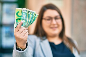 Young hispanic plus size businesswoman smiling happy holding israel shekels banknotes at the city.