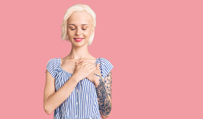 Young blonde woman with tattoo wearing casual clothes smiling with hands on chest with closed eyes...