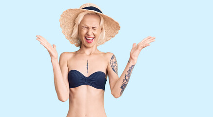 Young blonde woman with tattoo wearing bikini and summer hat celebrating mad and crazy for success with arms raised and closed eyes screaming excited. winner concept