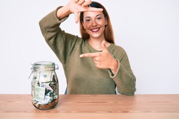 Young brunette woman sitting on the table with jar full of dollars smiling making frame with hands...