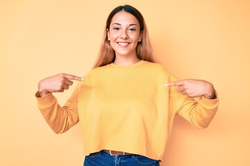 Young brunette woman over yellow background looking confident with smile on face, pointing oneself...