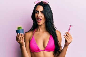 Beautiful hispanic woman wearing bikini holding cactus and razor angry and mad screaming frustrated...
