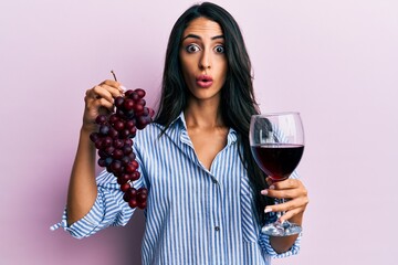 Beautiful hispanic woman holding branch of fresh grapes and red wine afraid and shocked with surprise and amazed expression, fear and excited face.