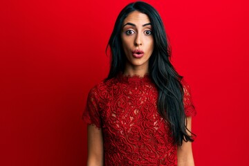 Beautiful hispanic woman wearing elegant clothes over red background scared and amazed with open...