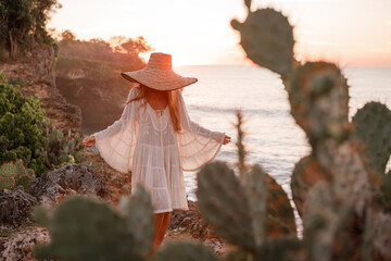 Unrecognizable stylish beautiful woman in white dress  and straw hat posing against  sea near green...