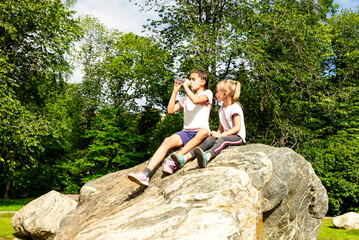 Two little girls sit on a large boulder in a city park and drink water from a bottle. © Ludmila