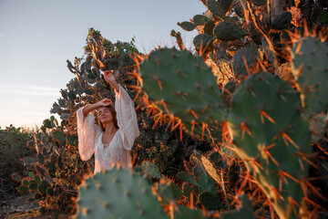 Young woman dressed in white dress and straw hat  standing among cactuses