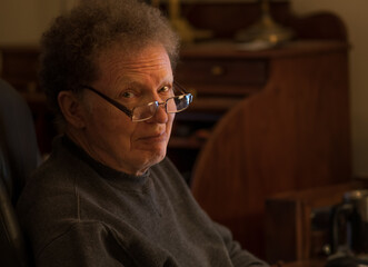 Fototapeta premium Senior man in glasses looking up from his dimply lit study room in evening; one side of his face is lit by table lamp; playful expression on his face