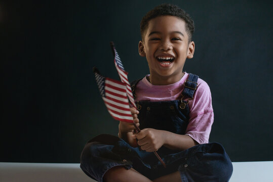 Portrait of Dark skinned child boy sit on table and waves the national American flag on black background, 4th of july, independent day