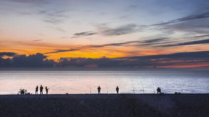 Evening twilight on the seashore. Silhouettes of fishermen, fishing rods, bicycles are visible. The sky is colored purple-orange. Black Sea. Russia. Sochi