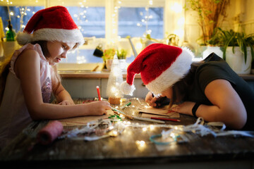 Caucasian girls children in santa red caps draw writing letters to Santa Claus at the table against the background of lights