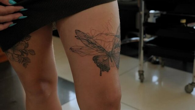 A young woman looks at her new tattoo on her leg in the mirror in a tattoo parlor. Image of a butterfly in black ink on the leg of a young woman.