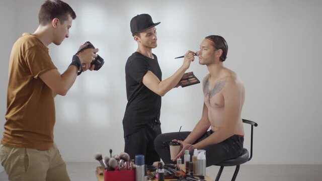 Medium shot of photographer taking photos of male beautician doing make-up on shirtless male client sitting on chair in studio