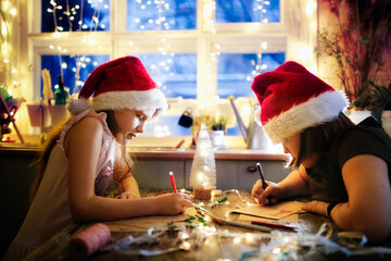 Caucasian girls children in santa red caps draw writing letters to Santa Claus at the table against the background of lights