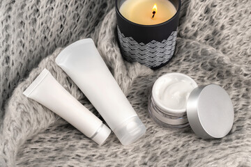 Fototapeta na wymiar Two white cosmetic tubes, a jar of cream, and a burning candle on a gray knitted fabric