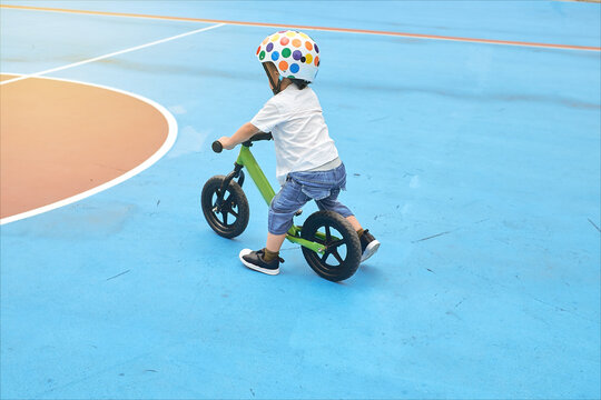 Backside Of Little Asian Toddler Boy Child Wearing Safety Helmet Learning To Ride His First Balance Bike, Kid Playing & Cycling At The Park