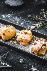 Homemade mini stollen with icing, dried cranberries and pistachios on a black tray on a black background, decorated with silver stars. Vertical stock photo.