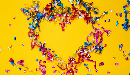 Colorful bright confetti and streamers in form of heart on yellow background, top view. Copy space for text. Holiday mood, Valentine day, Christmas, New year, birthday or funny party. Symbol of love