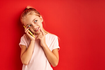 sweet child girl talking with someone on phone, looking away, flirting, listening to news by friend, chatting. she hold smartphone near ears, isolated on red background