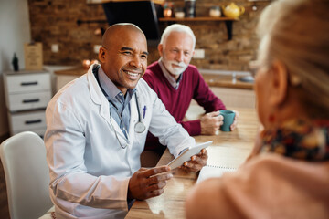 Happy black doctor using touchpad during a visit at mature couple's home.