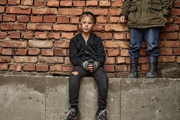 street kid boy sits on the concrete outdoors looking at camera, having no home, need shelter