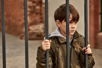 street boy stands behind bars, looks at people passing by, wants to live happily, depressed, need...