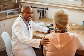 Happy black doctor talking to mature woman while visiting her at home.