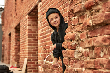 joyful street boy playing games using slingshot, lookin at side, hiding behind wall