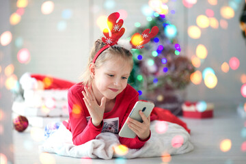 A little girl waves her hand and wishes a Happy New Year using a mobile phone for video calls to friends . Christmas online holiday remote celebration X mas  in lockdown coronavirus