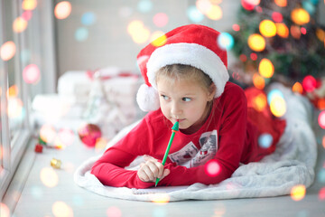 A smiling girl in a Santa hat writes a letter, dreams of gifts to Santa Claus. Child lying on a blanket on the floor of a house with christmas lights