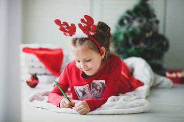 A smiling girl in a hoop on her head with deer antlers writes a letter, dreams of gifts to Santa Claus. Child lying on a blanket on the floor of a house with christmas lights