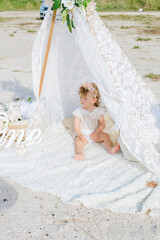 Fototapeta na wymiar A little cute girl with red curly hair in summer, on the seashore in an airy lace bodysuit and a floral headband in a lace tent, devoried with marine details, shells and cotton. 1st BIRTHDAY. 