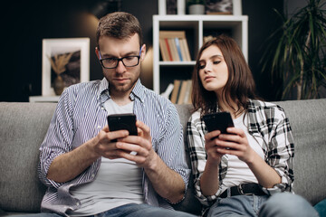 Young man and woman in casual clothing relaxing on grey couch and using modern smartphones. Happy people staying together at home with gadgets.
