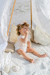Fototapeta na wymiar A little cute girl with red curly hair in summer, on the seashore in an airy lace bodysuit and a floral headband in a lace tent, devoried with marine details, shells and cotton. 1st BIRTHDAY. 
