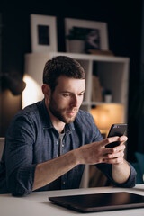 Attractive young man in denim shirt using smartphone while spending free time at home. Concept of relaxation and modern technology.